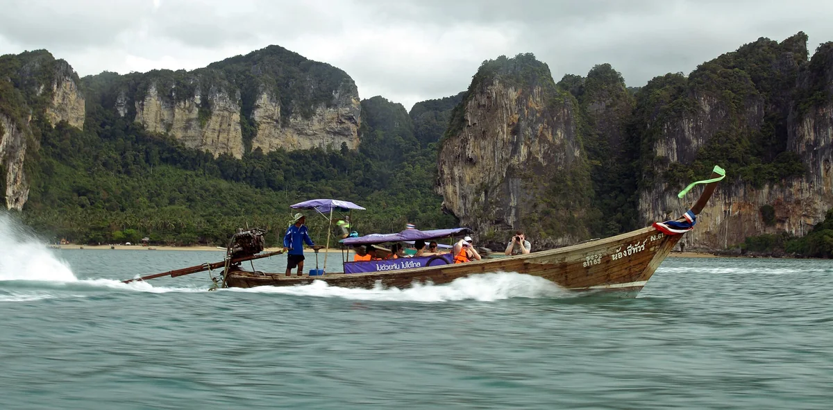 A longtail boat crossing the water with Tonsai Beach and the limestone cliffs of the Railay peninsula behind it